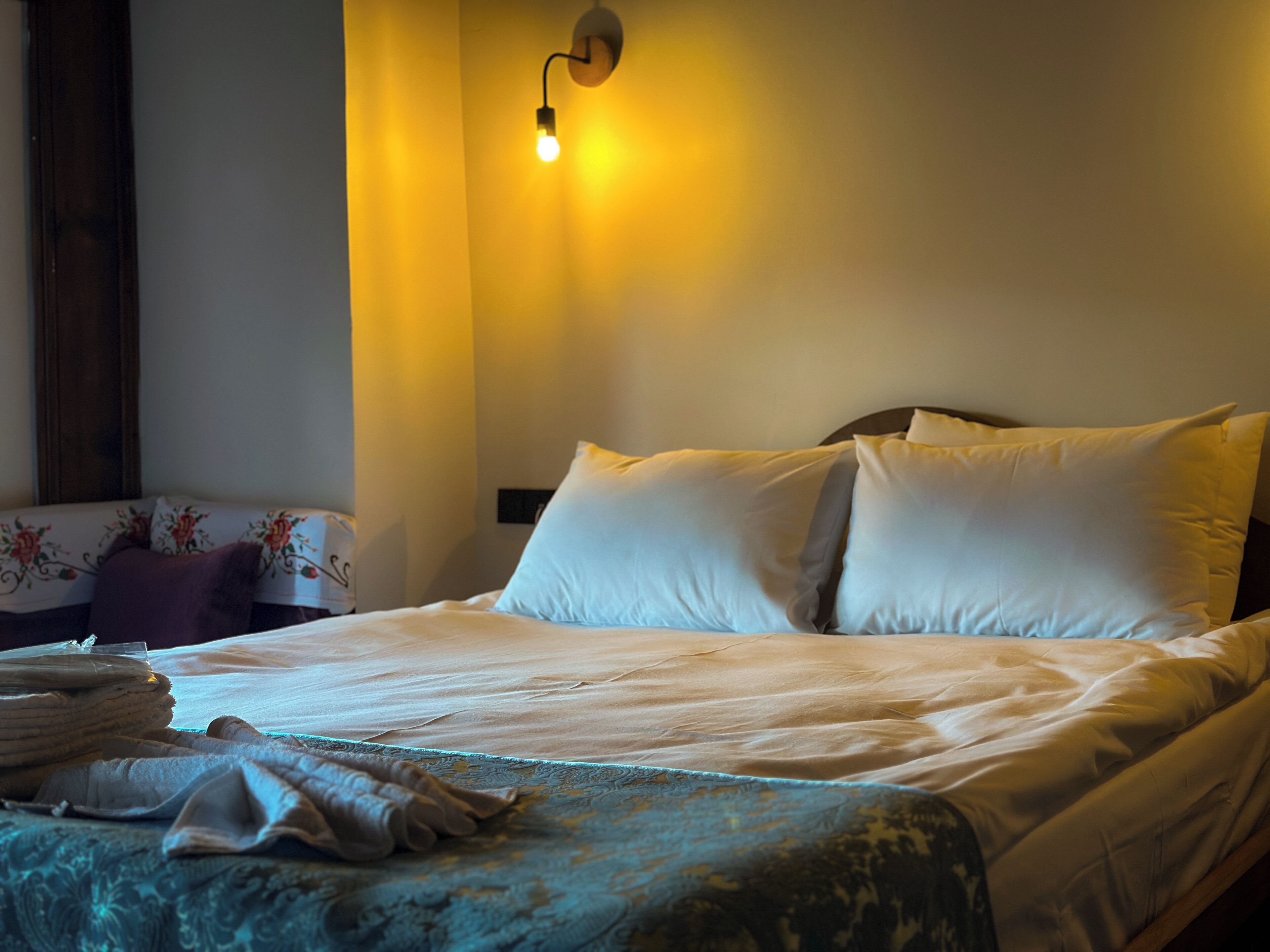 Una cama bien hecha con sábanas blancas y una manta estampada en azul, un aplique de pared y un cojín con motivos florales sobre una mesita de noche.