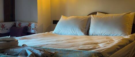 Una cama bien hecha con sábanas blancas y una manta estampada en azul, un aplique de pared y un cojín con motivos florales sobre una mesita de noche.