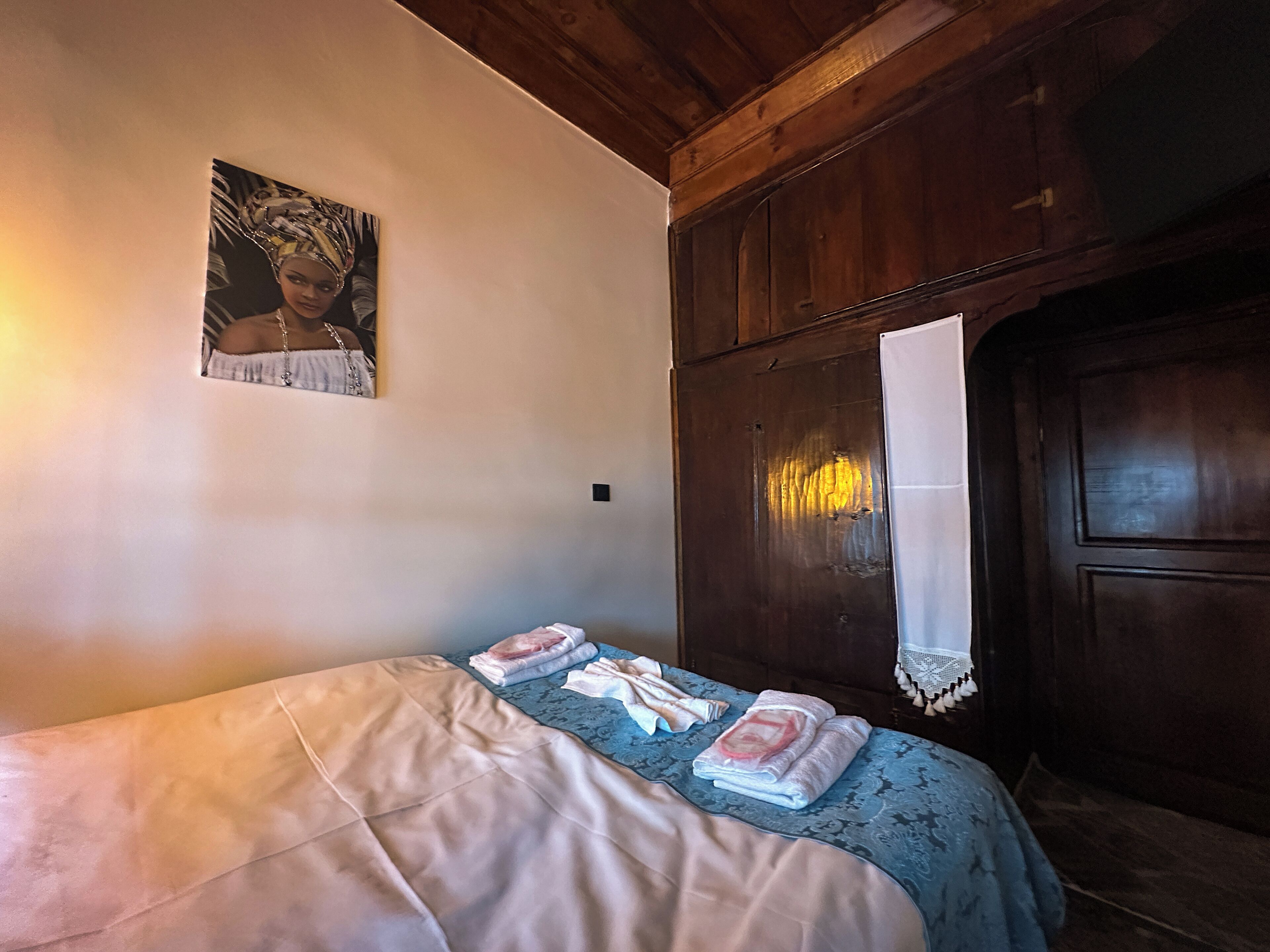 Une chambre à coucher avec un lit, un tableau accroché au mur, une armoire en bois et une fenêtre ornée d’un rideau blanc.