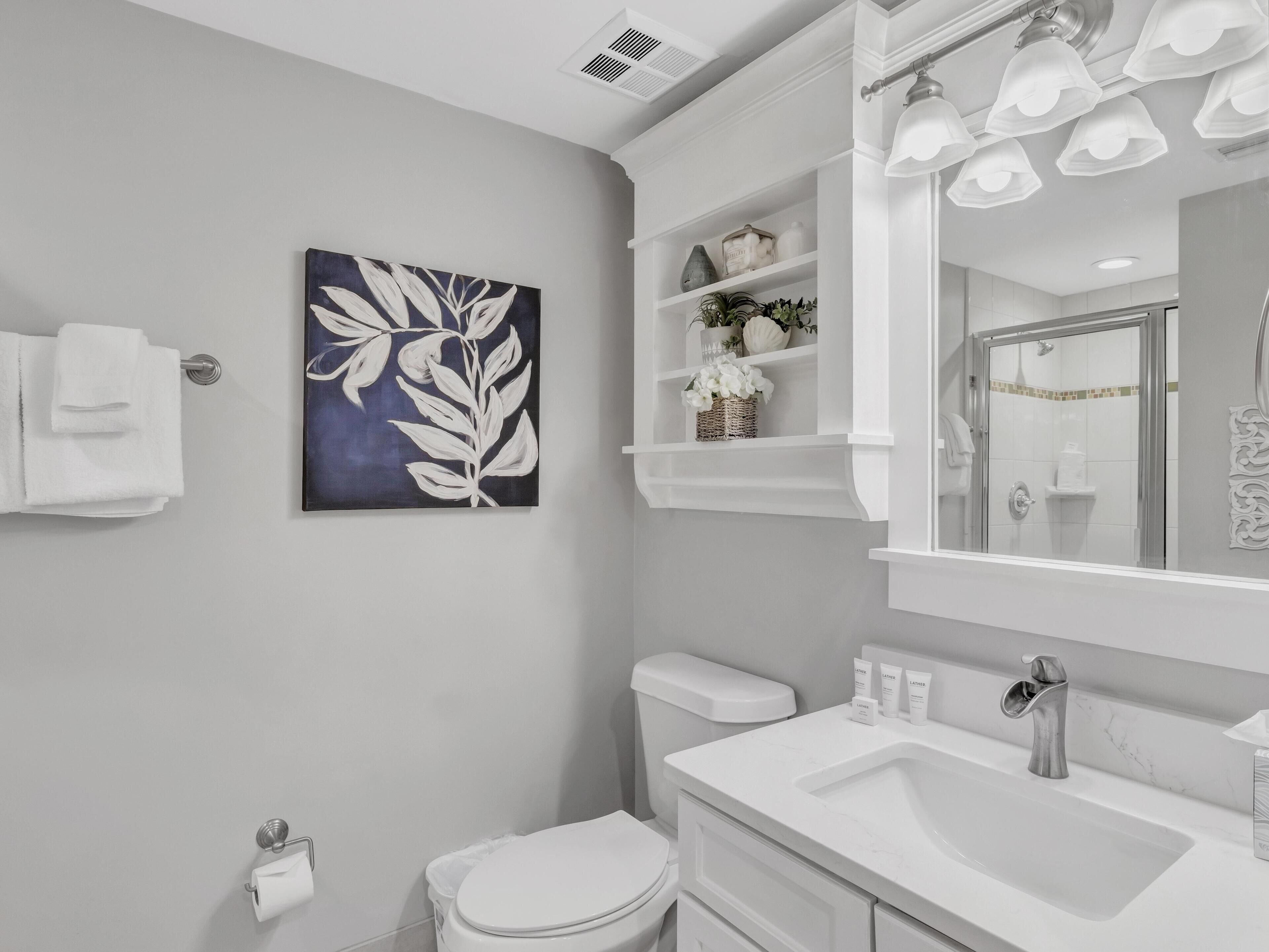 A bathroom with a white sink, toilet, and a large mirror with lights. There is a towel rack and a painting on the wall.