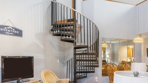 A modern living room with a curved black staircase, a flat-screen TV, wicker furniture, and a sign that reads "Adventure Awaits". - Fairway Haven For 9! (Branson)