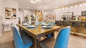 Un espace repas moderne avec une table en bois dressée pour quatre personnes, des chaises à l’assise bleue, et une cuisine en arrière-plan équipée d’appareils électroménagers en acier inoxydable.