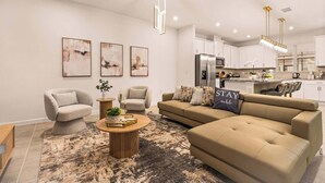 Un salon moderne avec un canapé beige, deux fauteuils, une table basse et un tapis. Trois tableaux abstraits ornent le mur, et on aperçoit une cuisine au fond, ainsi qu'un lustre.