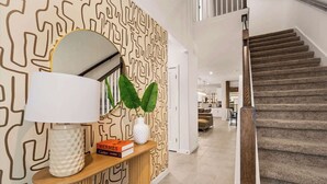 Un intérieur moderne avec un mur orné d’un motif, une console en bois avec un miroir, des livres et un vase contenant des feuilles vertes.