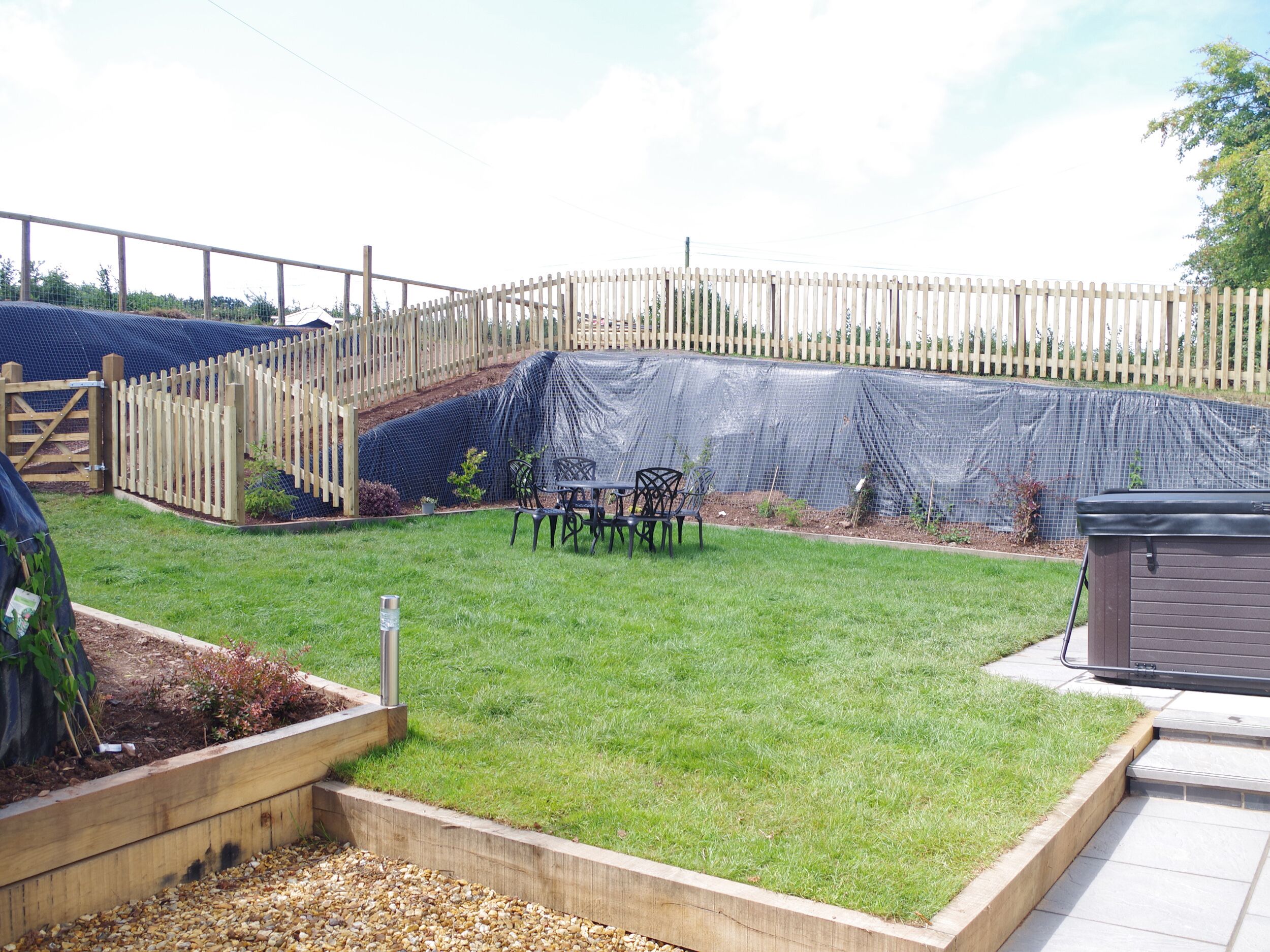 Un patio trasero cercado con un área verde, una terraza de madera y una pequeña mesa con sillas.