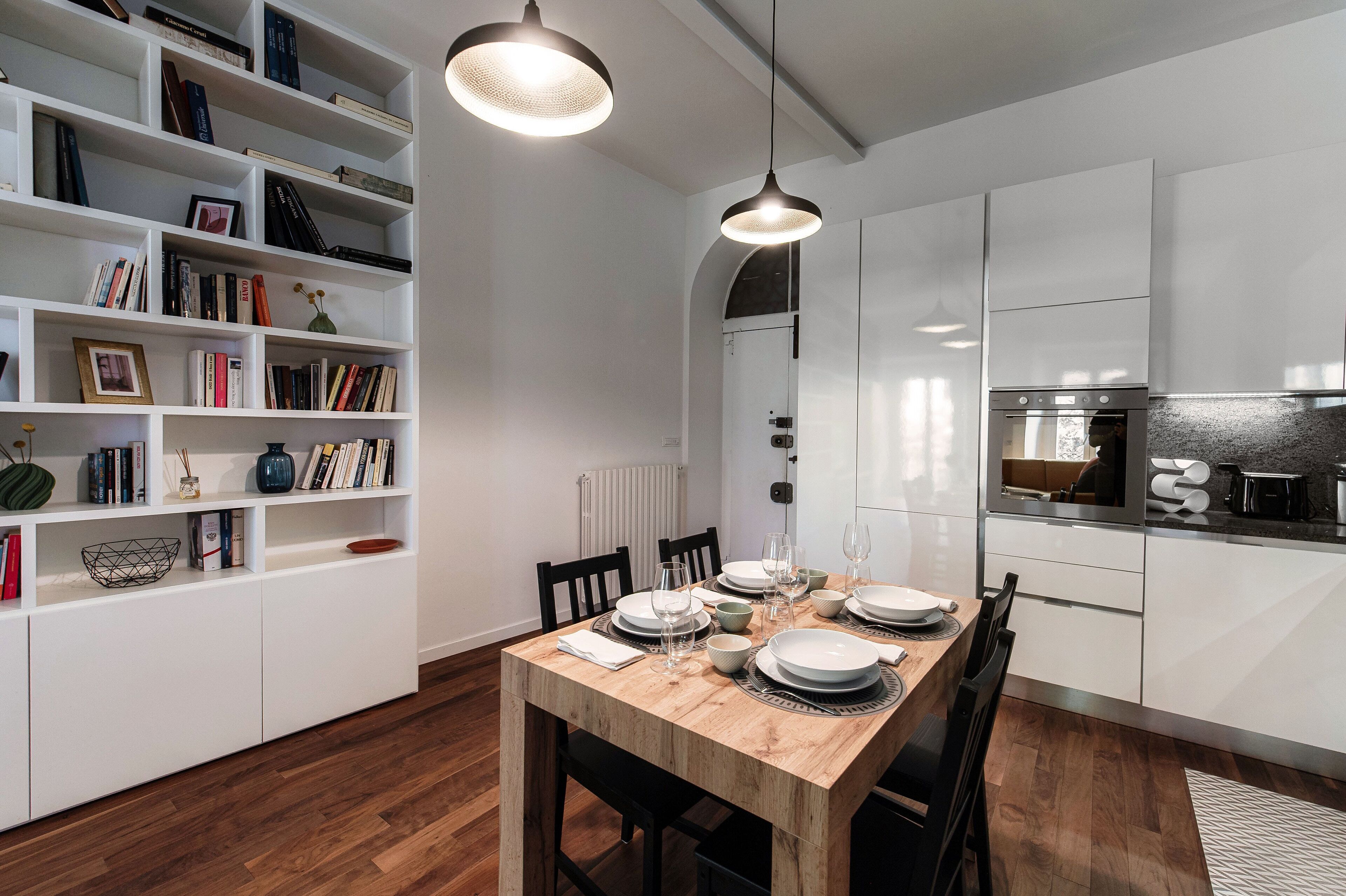 Een moderne keuken met witte kasten, een houten eettafel voor vier personen, en een boekenkast vol boeken en decoratieve objecten.