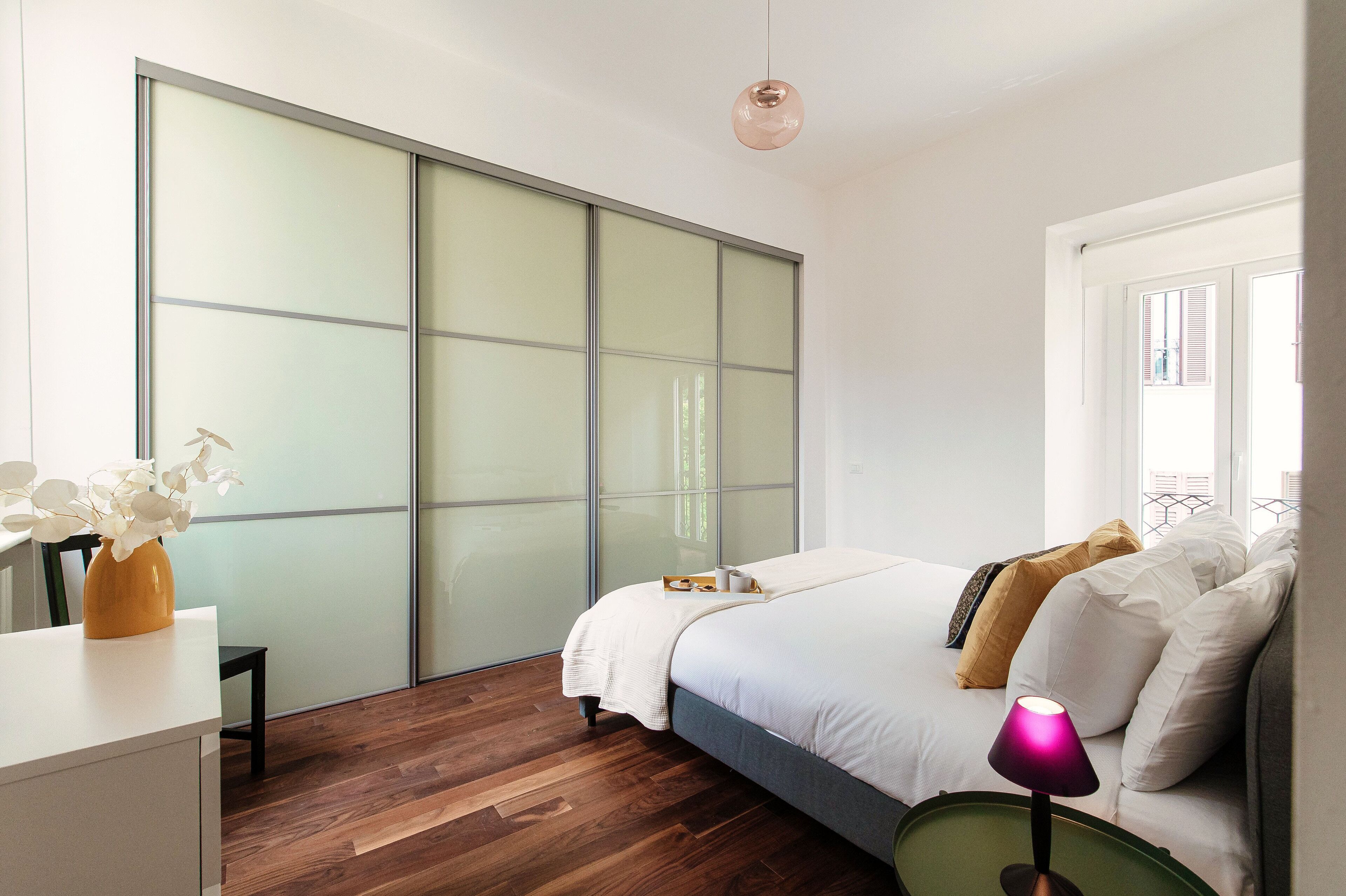 Um quarto moderno com uma grande porta de vidro deslizante, uma cama com roupa de cama branca, uma mesa de cabeceira com abajur e um vaso de flores brancas.