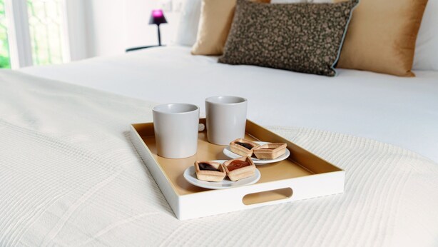 Un lit bien fait, avec un plateau de viennoiseries et deux tasses posées sur un couvre-lit blanc.