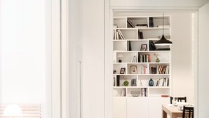 Une pièce moderne et bien éclairée, avec une bibliothèque blanche remplie de livres et d’objets décoratifs, un coin repas doté d’une table et de chaises en bois, et un plancher en bois.