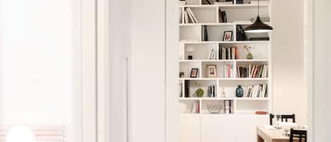 A modern, well-lit room with a white bookshelf filled with books and decorative items, a dining area with a wooden table and chairs, and a wooden floor.