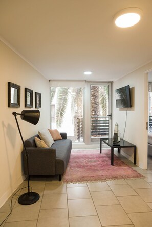 A living room with a sofa, a floor lamp, a TV, and a view of palm trees through the glass door. - Central 1BR1BA in Santiago Balcony & Nearby Metro (Unknown)