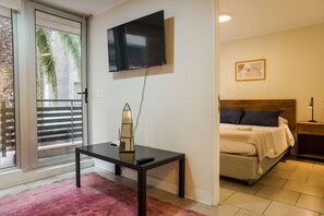 A hotel room with a bed, a TV mounted on the wall, a small table with a remote control, and a window with a view of palm trees.