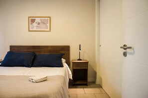 A hotel room with a bed, pillows, a bedside table with a lamp, a framed picture on the wall, and a door.
