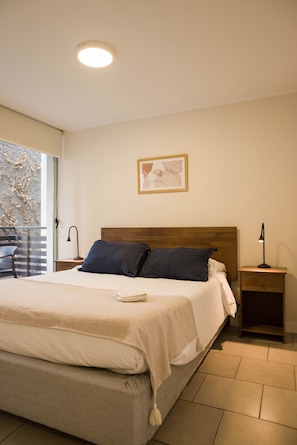 A bedroom with a bed, a nightstand, a lamp, a framed picture on the wall, and a window with a view of foliage. - Central 1BR1BA in Santiago Balcony & Nearby Metro (Unknown)