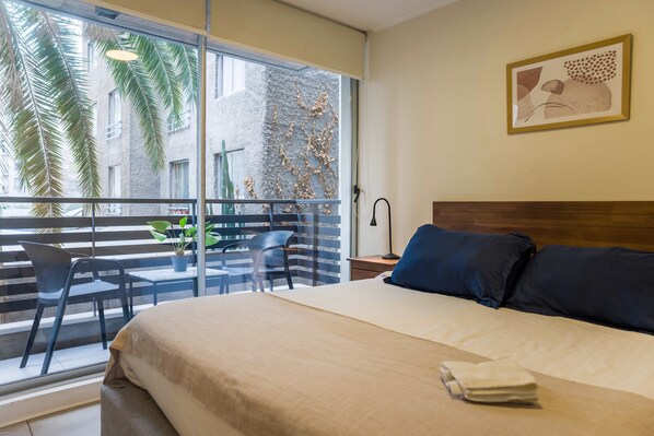 A hotel room with a large bed, a balcony with chairs and plants, and a view of palm trees and buildings. - Central 1BR1BA in Santiago Balcony & Nearby Metro (Unknown)
