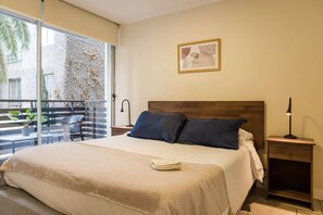 Une chambre d’hôtel avec un grand lit, une table de chevet avec une lampe, une commode et une vue sur un balcon agrémenté de plantes.