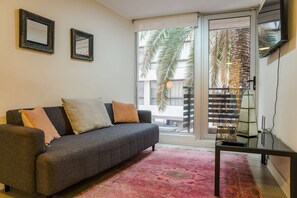 A living room with a grey sofa, two framed pictures, a pink rug, and a sliding glass door leading to a balcony with a palm tree.