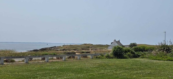Un paysage côtier avec une zone herbeuse, un petit bâtiment et une étendue d’eau en arrière-plan.