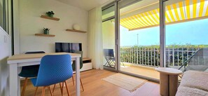 Un salon moderne avec un plancher en bois, un fauteuil bleu, un bureau blanc et un téléviseur. Il y a un balcon avec un auvent jaune à rayures et une vue sur la verdure.