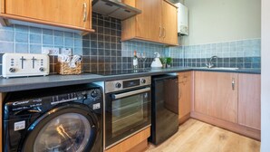 Een compacte keuken met houten kastjes, een inbouwoven, een vaatwasser en een wasmachine.