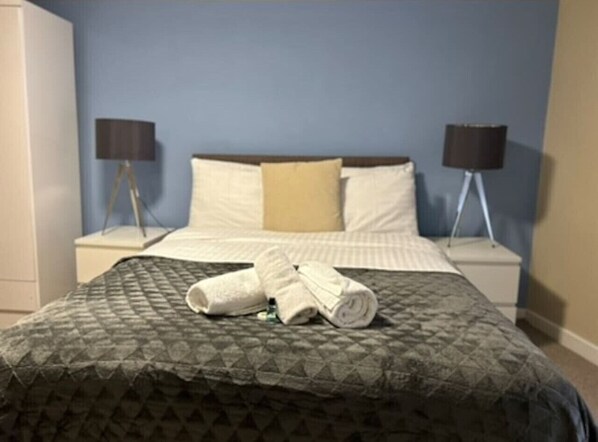 A neatly made bed with a patterned bedspread and two rolled towels. There are two bedside lamps on either side of the bed.
