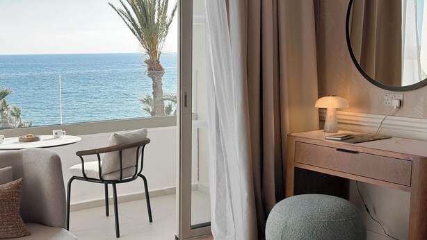 Une chambre d’hôtel avec un balcon, un bureau, une chaise, un pouf rond et une vue sur la mer.