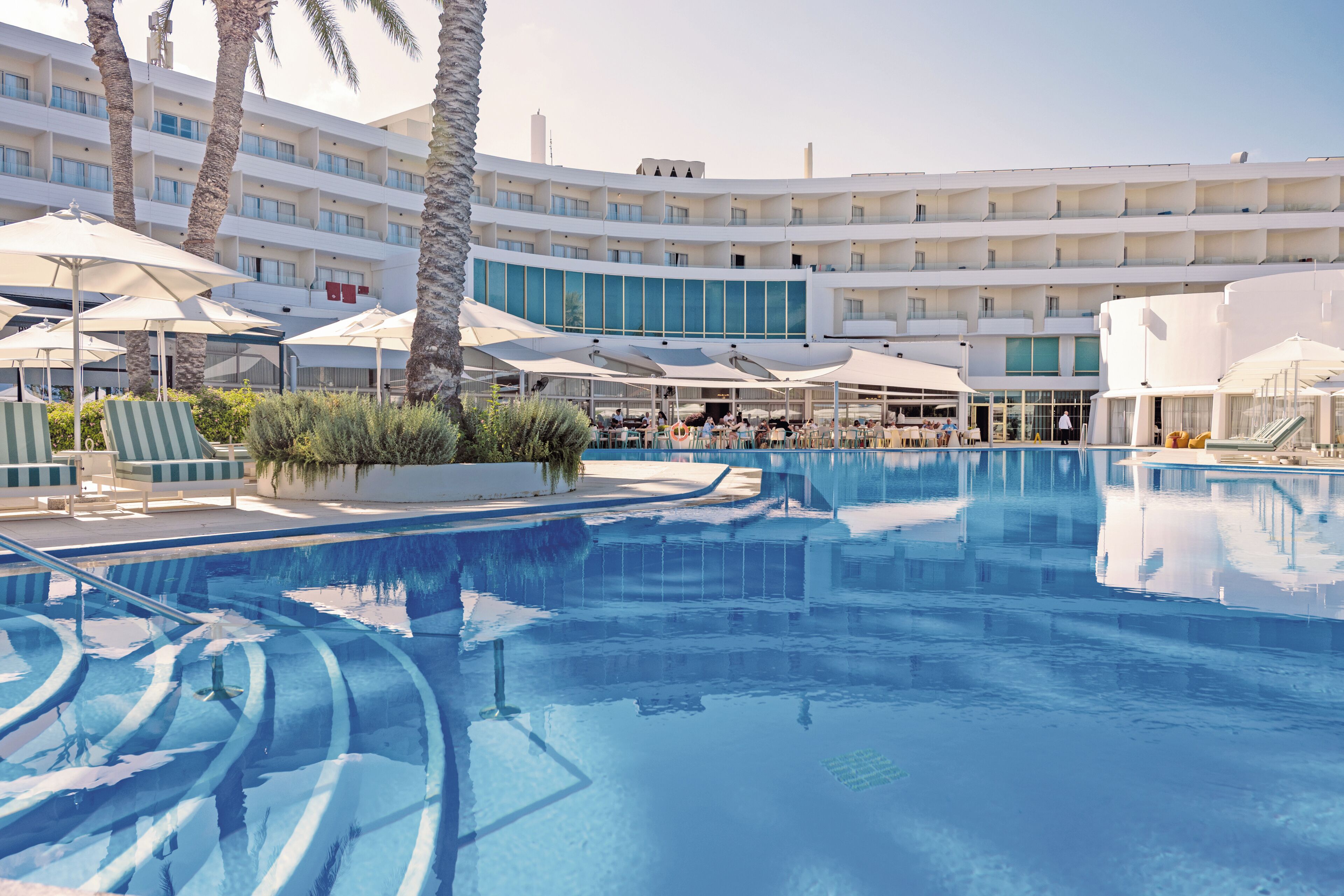 A hotel pool area with multiple levels of balconies, sun umbrellas, and lounge chairs.