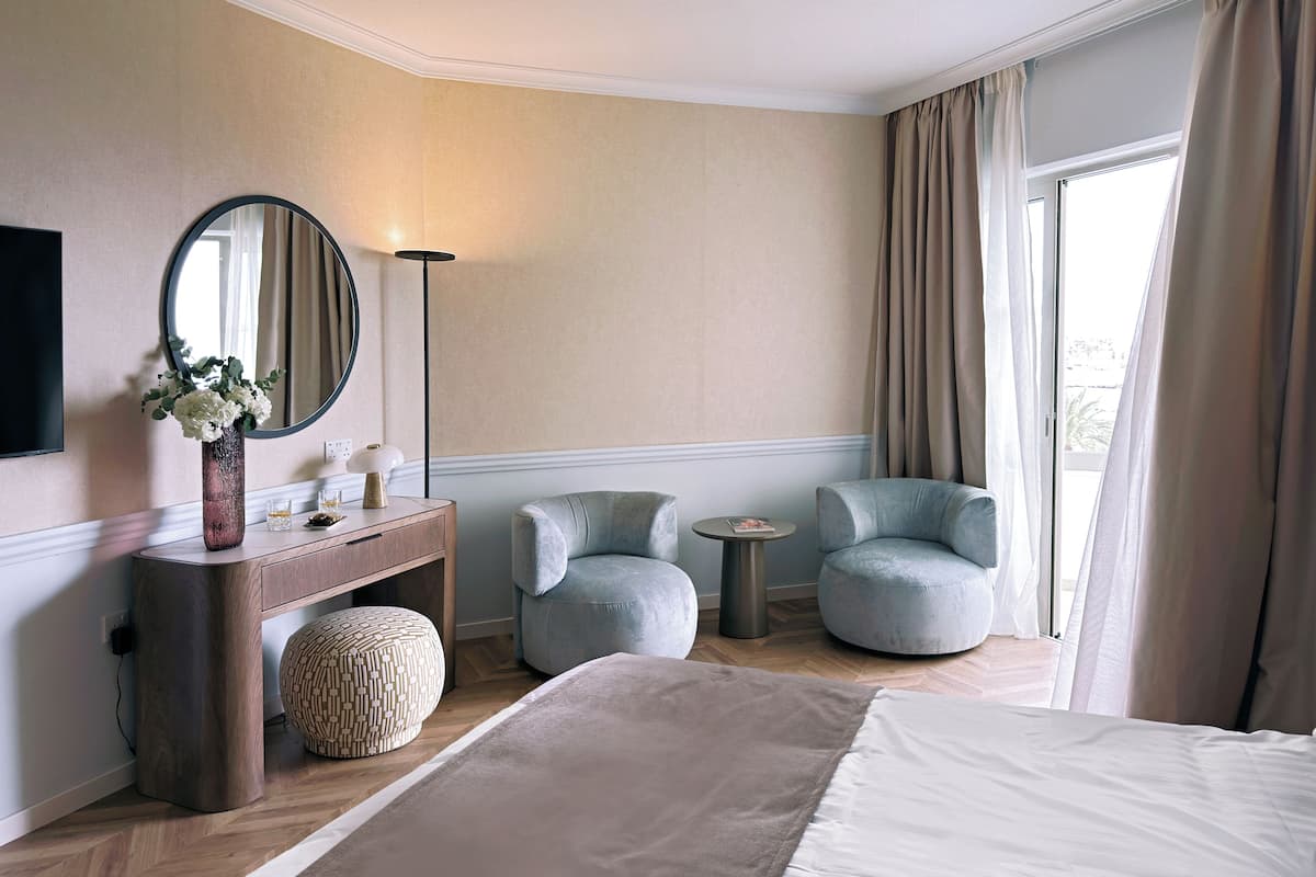 Quarto de hotel com uma cama, um espelho redondo, uma mesa consola de madeira, duas poltronas, uma pequena mesa redonda e uma grande janela com cortinas.