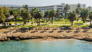 Un complexe hôtelier doté d’une plage, d’une eau cristalline, de palmiers et d’un grand bâtiment comportant de nombreux balcons.