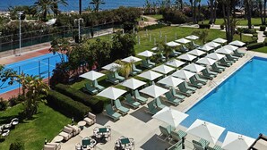 Un espace piscine aménagé dans un complexe hôtelier, avec des chaises longues et des parasols, un court de tennis et une vue sur l’océan.