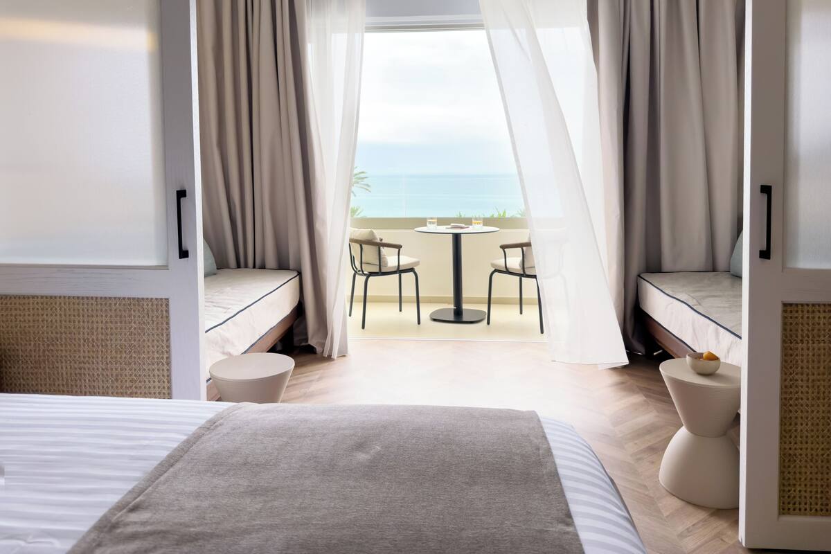 Quarto de hotel com uma cama, uma mesinha, duas cadeiras e vista para o mar.