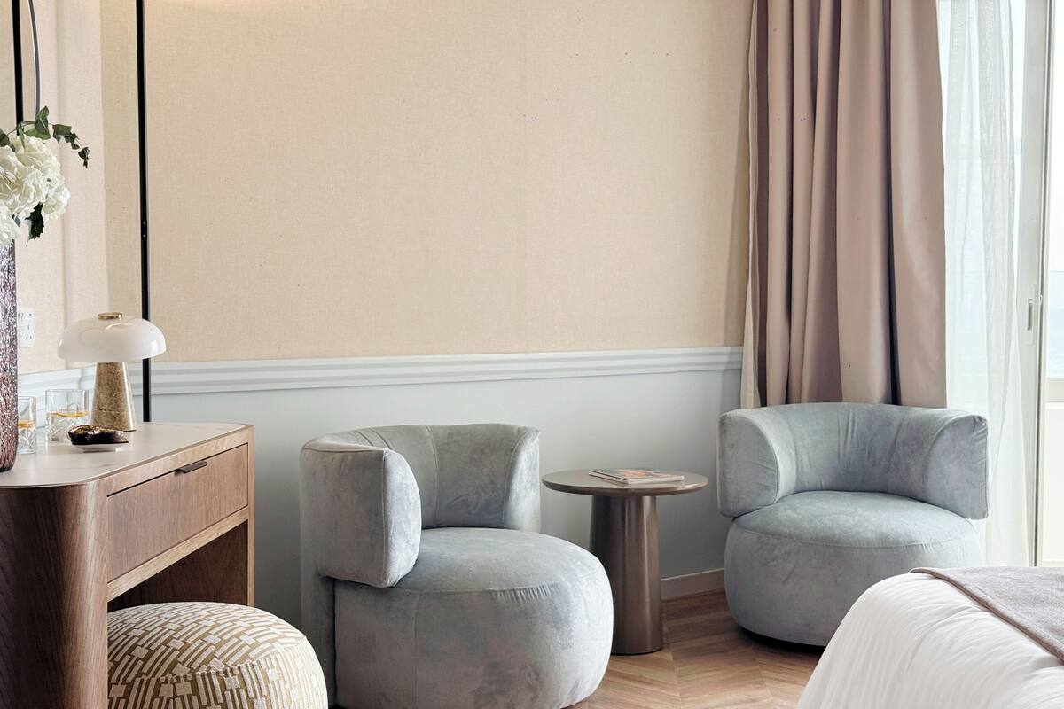 Quarto de hotel moderno com cómoda de madeira, duas poltronas cinzentas, uma mesa redonda pequena e uma cama grande.