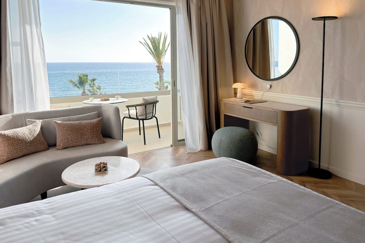 Quarto de hotel com cama, escrivaninha, cadeira, espelho redondo e vista para o mar.