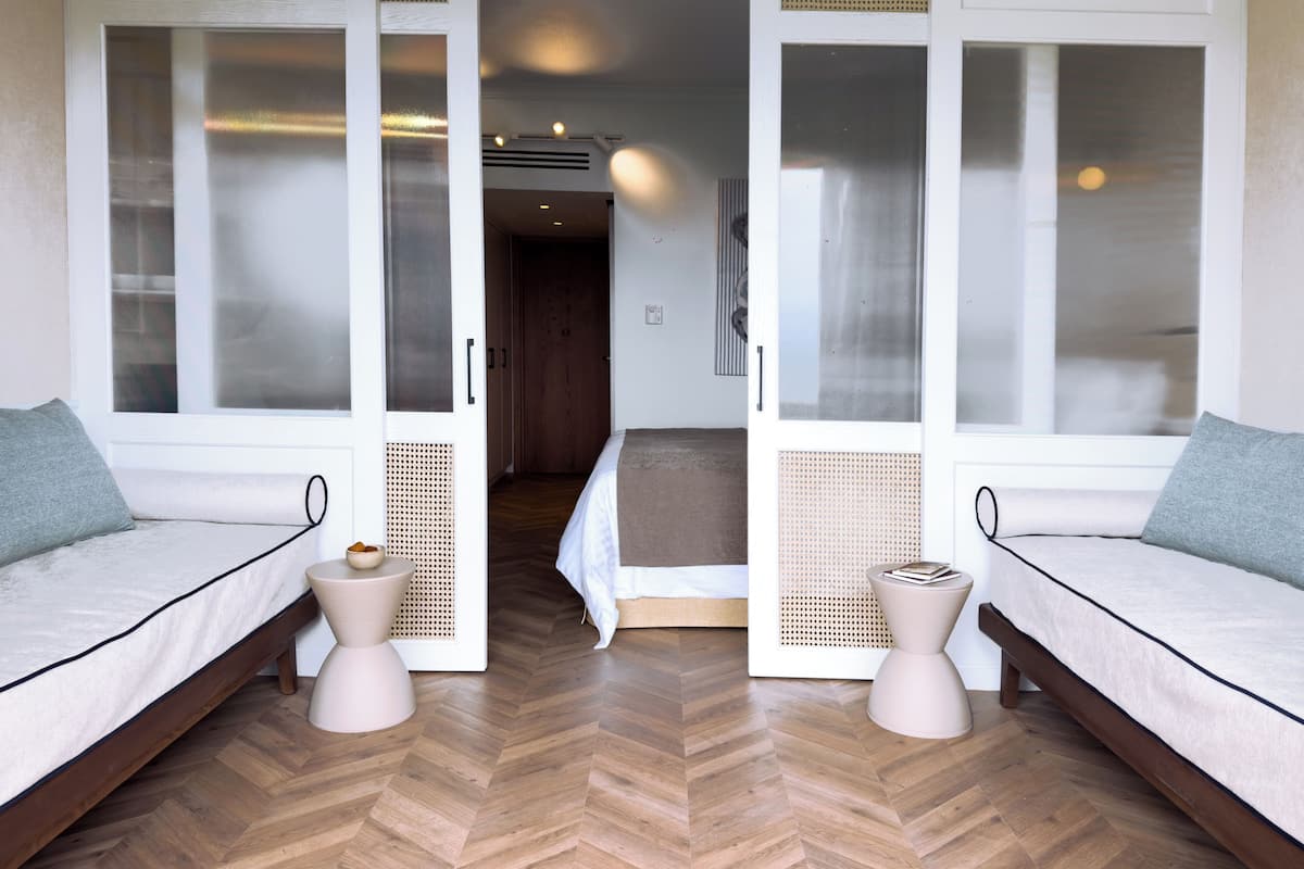 Quarto com duas camas, piso de madeira em padrão espinha de peixe e porta que dá para um corredor.