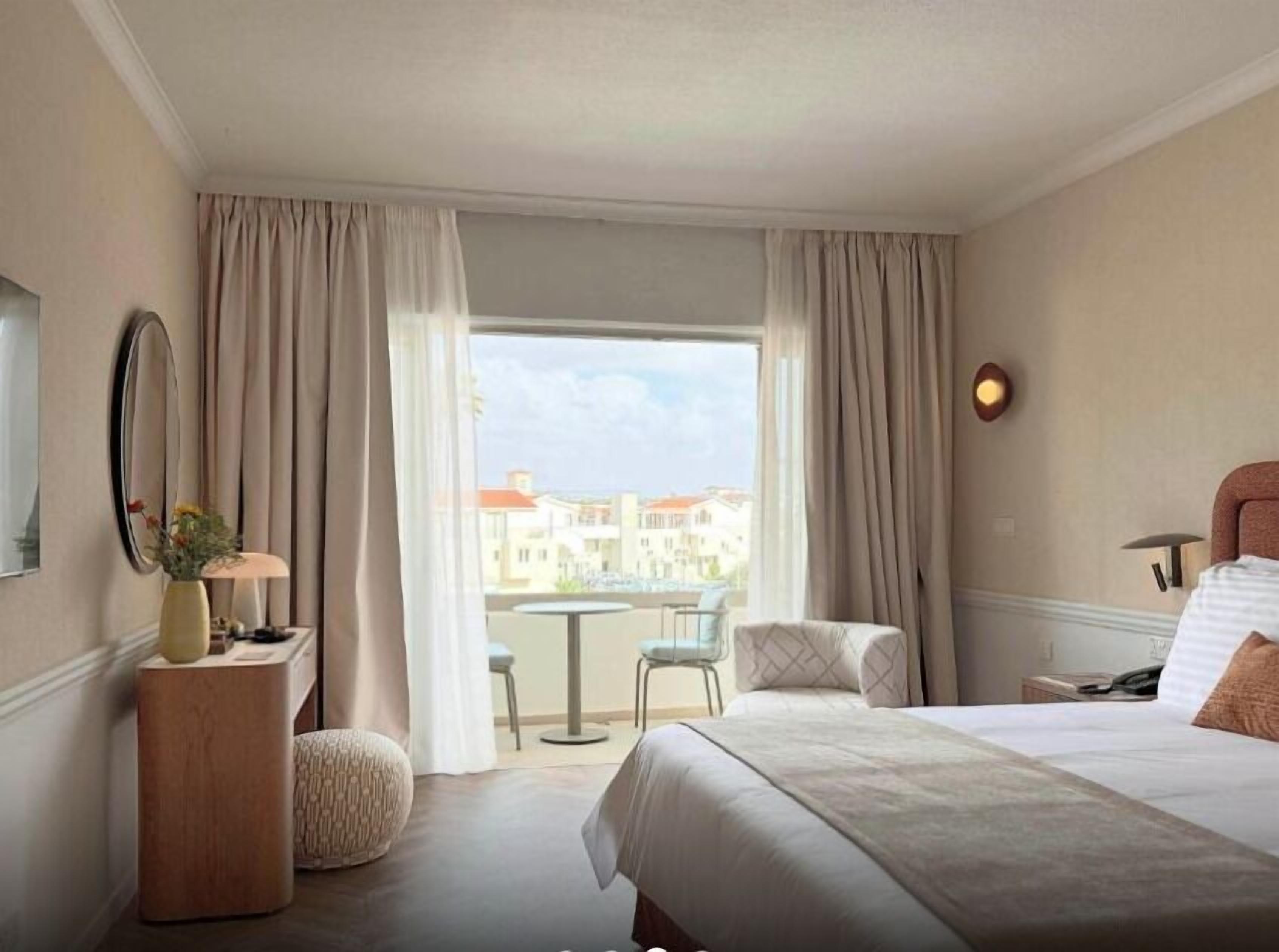 Ein Hotelzimmer mit Bett, Schreibtisch, Stuhl, rundem Tisch und Blick auf Gebäude durch große Fenster.