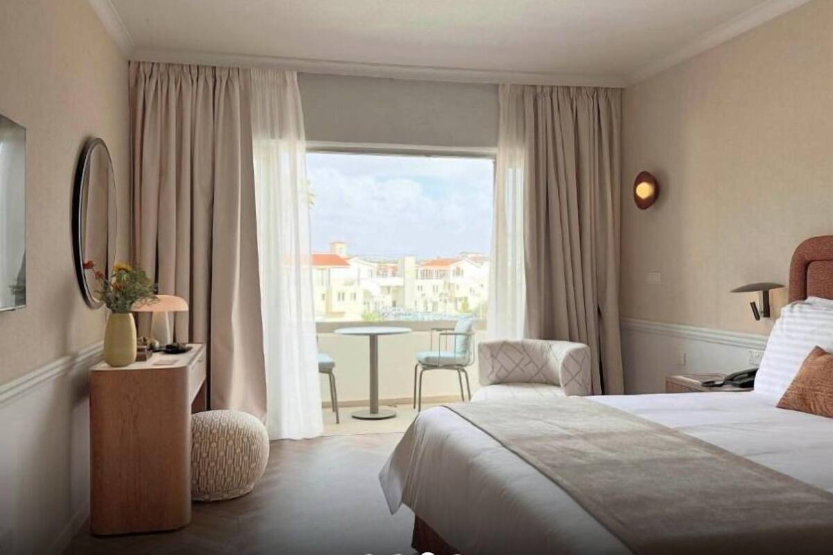 Ein Hotelzimmer mit Bett, Schreibtisch, Stuhl, rundem Tisch und Blick auf Gebäude durch große Fenster.