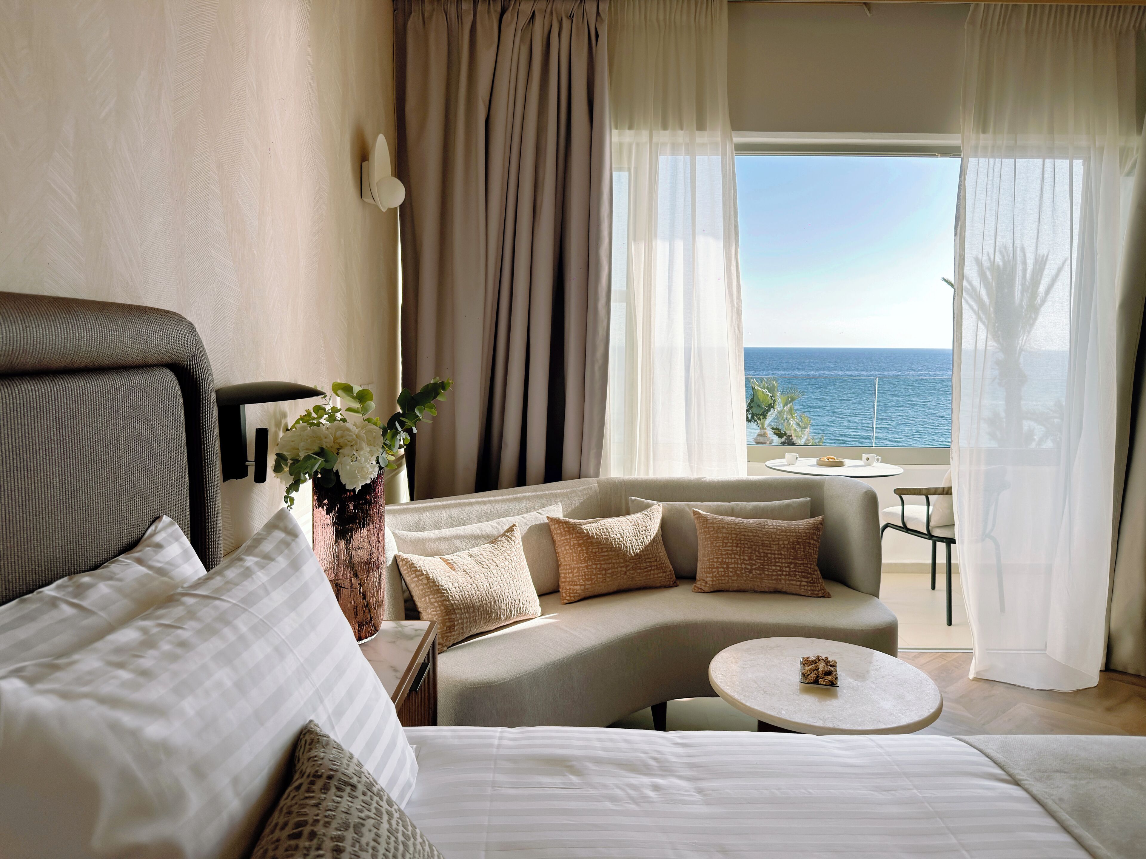 A hotel room with a bed, a sofa, a small table, a vase with flowers, and a view of the sea.