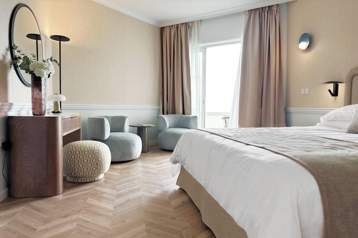 Quarto de hotel com uma cama grande, duas poltronas, uma mesinha, uma cômoda e um espelho.