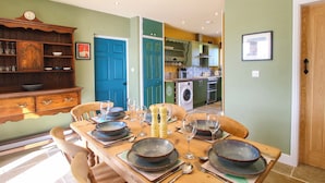A dining area with a wooden table set for six, surrounded by chairs. A kitchen with green cabinets and a blue door is visible in the background. - Dingle (Kidderminster)