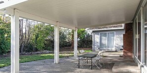 A covered patio with a table and chairs, a brick building in the background, and a grassy area with trees. - Family, Pet & Long Vacation Friendly, Fenced Yard (Sacramento)