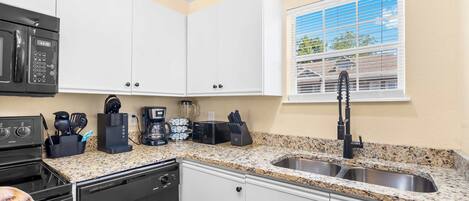 Una cocina con gabinetes blancos, encimera de granito, pileta de acero inoxidable, microondas negro, cafetera y ventana con persianas.