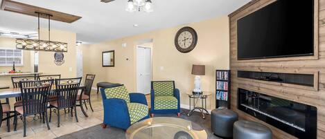 A living room with a glass coffee table, blue and green patterned chairs, a wooden TV stand, and a ceiling fan.