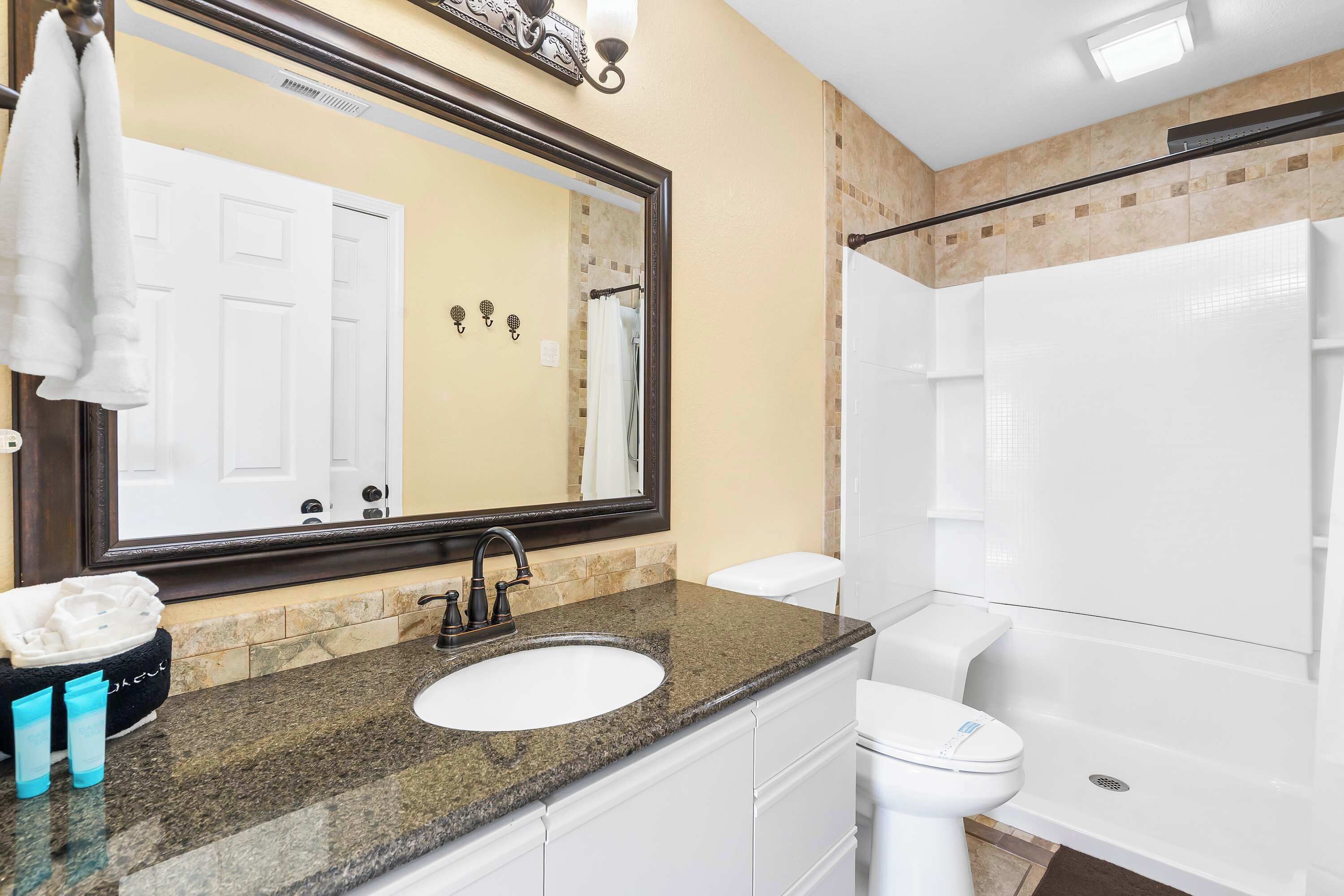 A bathroom with a granite countertop sink, a white toilet, a large mirror, and a shower area.
