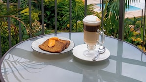 A table with a glass of coffee and toast on a balcony overlooking a pool and greenery. - Victoria Falls Oasis Hotel (Victoria Falls)