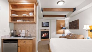 A compact hotel room with a kitchenette, a fireplace, a bed, and a small bedside table with a lamp.