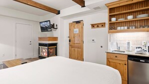 A cozy room with a fireplace, wooden beams, a TV mounted on the wall, and a kitchen area with wooden shelves and a dishwasher.