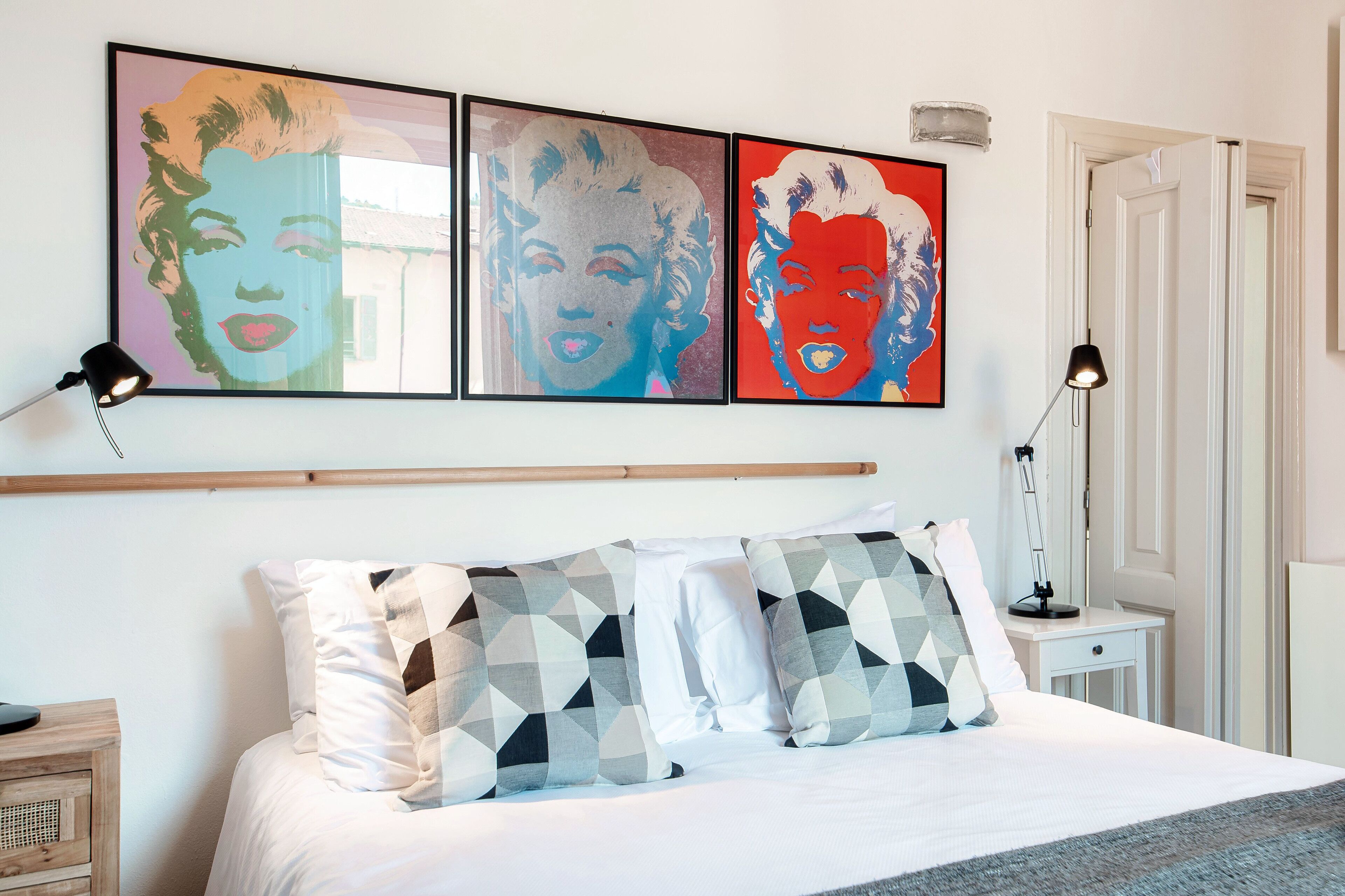 Un lit bien fait, agrémenté d’oreillers à motifs, une table de chevet avec une lampe, et trois œuvres d’art encadrées accrochées au mur au-dessus du lit.