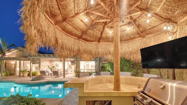 An outdoor pool area with a thatched umbrella, a built-in grill, and a flat-screen TV.