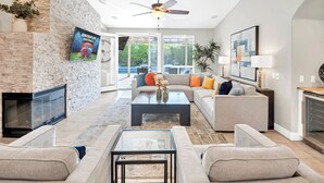 A modern living room with a fireplace, a ceiling fan, and a large window offering a view of the outdoors.