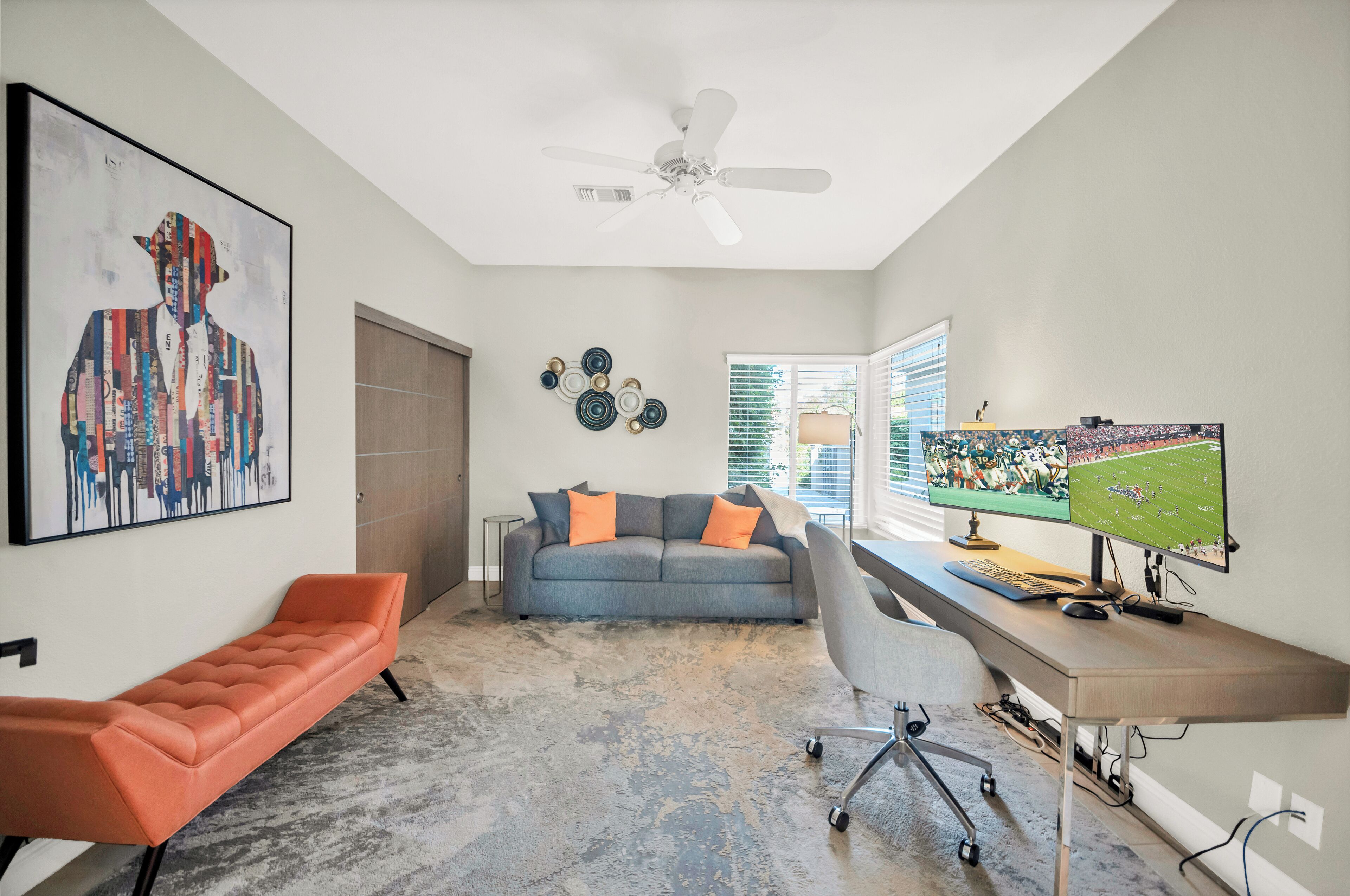 A modern living room with a gray sofa, orange bench, and a desk with a computer monitor.